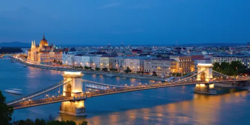 A panoramic view of a city at dusk, featuring a bridge over a river and illuminated buildings along the waterfront.