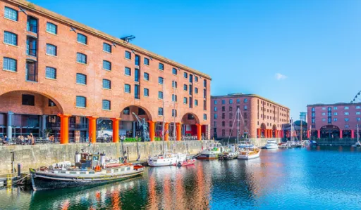 Historic red brick buildings line a waterfront, with boats moored in the calm water under a clear blue sky.