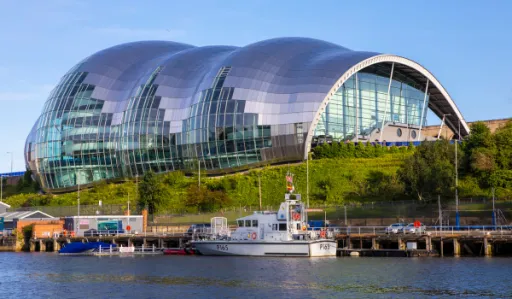 Modern building with a curved glass facade overlooks a river with boats docked nearby.