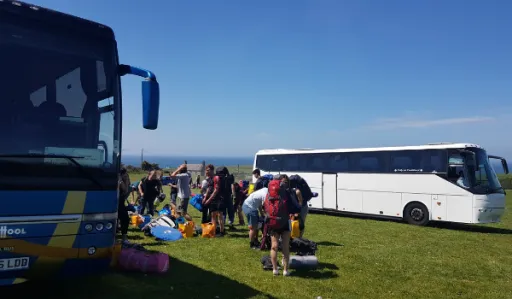 Two buses are parked on grass with people unloading bags and equipment under a clear blue sky.