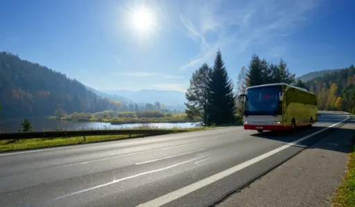 Yellow bus drives along a road beside a lake, with mountains and trees in the background under a sunny sky.