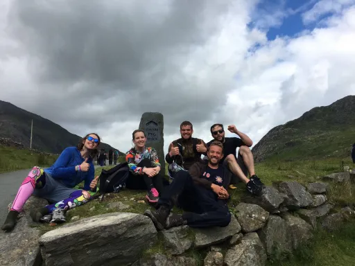 Five people sit on a stone wall near a sign for the Pyg Track, with mountains and cloudy skies in the background.