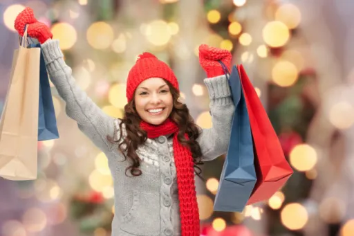 Person in a red hat and scarf holds shopping bags and smiles against a blurred festive background.