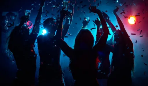 Silhouettes of people dancing with drinks under colourful lights and confetti.