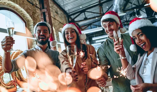 Group of people wearing Santa hats celebrate with drinks and sparklers in a festive setting.