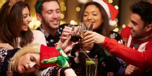 A group of people at a festive gathering, raising glasses in a toast, with one person resting their head on the table.