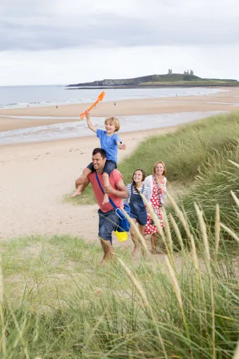 A person carries a child on their shoulders while two others walk behind them on a sandy beach with grass.