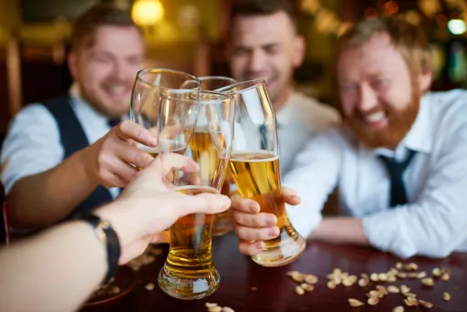 Four people clink large glasses of beer together at a table, with snacks scattered on the surface.