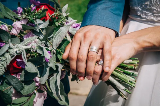 Two hands holding a bouquet of flowers, with wedding rings visible on both fingers.