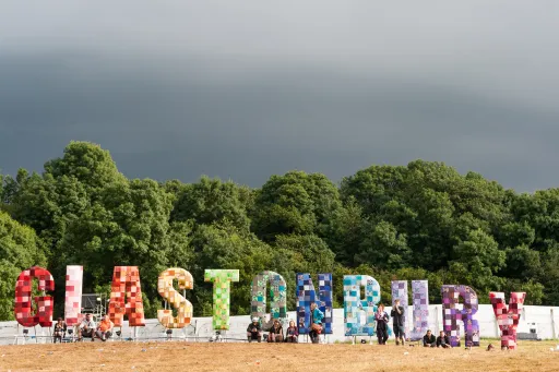 Colourful letters spelling GLASTONBURY in a field, with people sitting nearby and trees in the background.