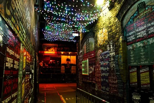 A narrow alleyway at night, featuring colourful lights above and vibrant murals on the brick walls.