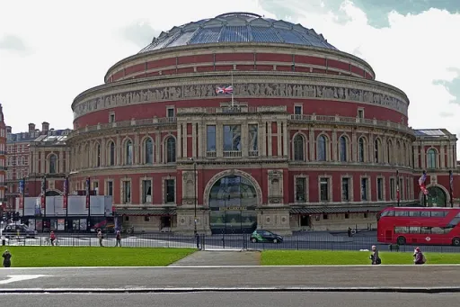 The image shows the exterior of a large circular building with a glass dome, surrounded by green grass and a red bus nearby.