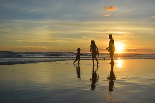 Silhouettes of three people walking on a beach at sunset, with reflections on the wet sand and waves in the background.