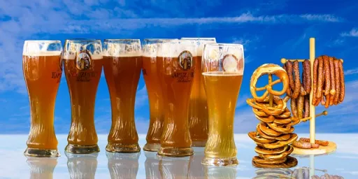 A row of six glasses of beer with a selection of pretzels and sausages on a stand against a blue sky.