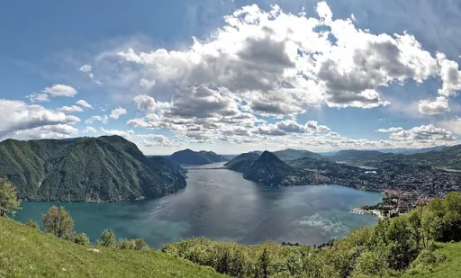 Panoramic view of a lake surrounded by mountains and hills under a cloudy sky.