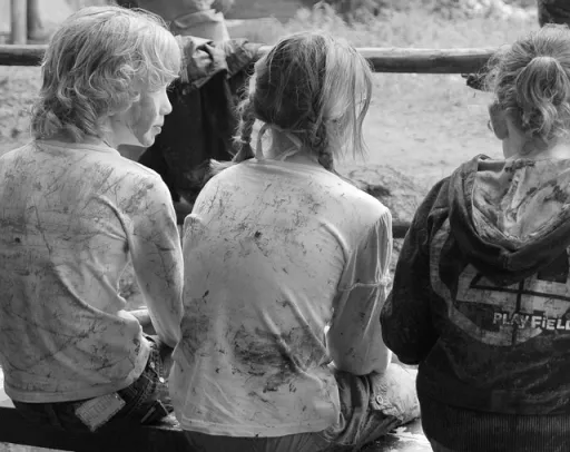 Three people sitting on a bench, viewed from behind, wearing dirty, light-coloured shirts in a natural setting.