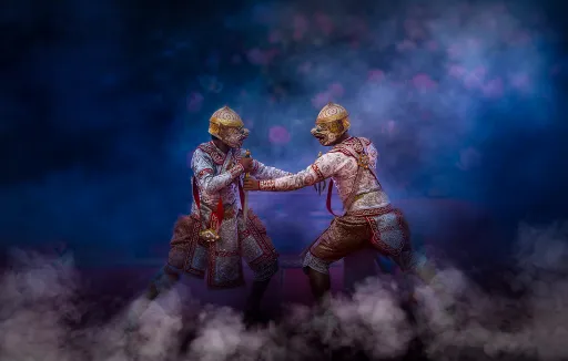 Two figures in ornate costumes and masks engage in a dramatic pose, surrounded by mist and a dark background.