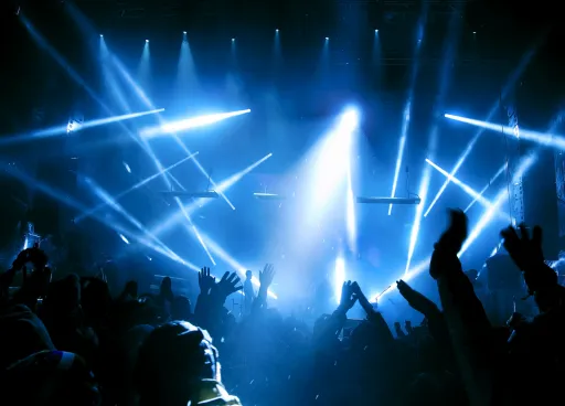 Crowd with raised hands at a concert, illuminated by blue and white stage lights in a smoky atmosphere.